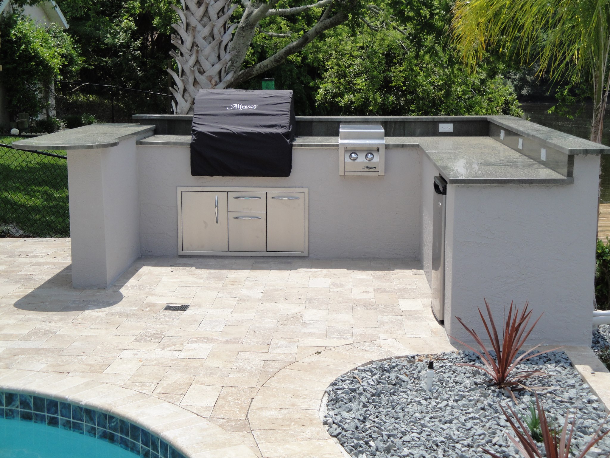 Pool-side U-shaped summer kitchen with travertine pavers, built-in Alfresco grill, stainless cabinets, and beverage fridge cut-out