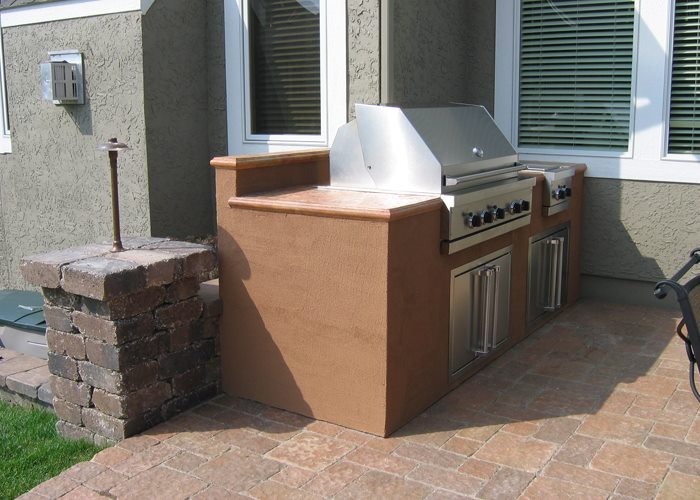 Stucco-front summer kitchen with copper-stained countertop, built-in stainless grill, and stone pillar accent on a paver patio